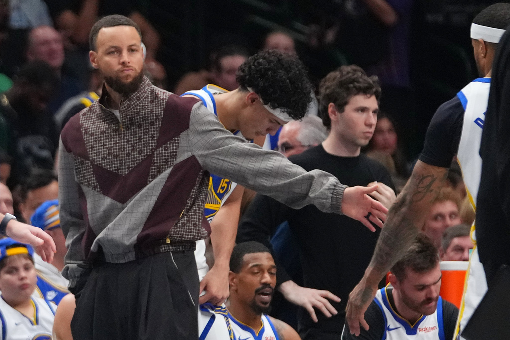 Golden State Warriors guard Stephen Curry wears street clothes while standing near his team's bench during a timeout against the Dallas Mavericks in the first half of an NBA basketball game Monday, March 23, 2026, in Dallas. (AP Photo/Julio Cortez)