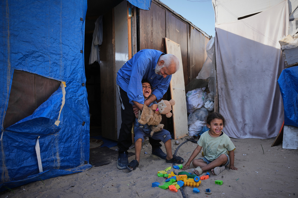 Mahmoud Nofal, 64, plays with his grandchildren, Tamim and Ranin Nofal, whom he has raised since their parents were killed in an Israeli airstrike, outside their tent in Khan Younis, southern Gaza Strip, June 2, 2026. (AP Photo/Abdel Kareem Hana)