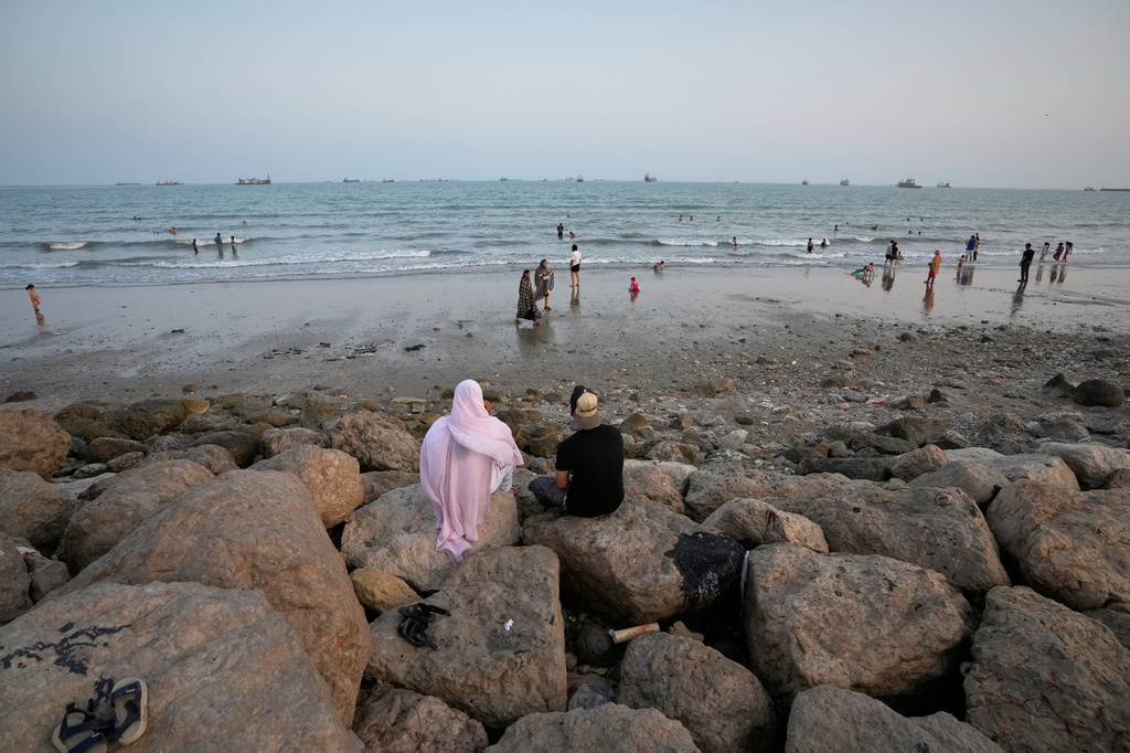 Two people sit on rocks overlooking a beach as others swim and commercial vessels lie anchored in the Strait of Hormuz off Bandar Abbas, Iran, Friday, Sept. 4, 2026. (AP Photo/Vahid Salemi)