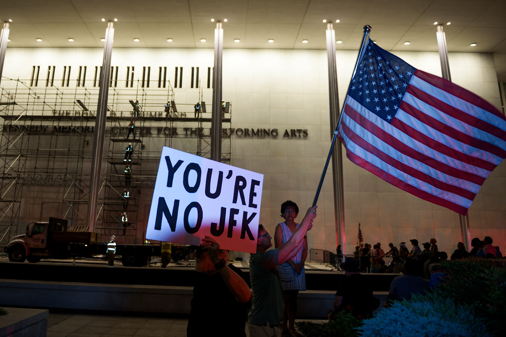 Protestors wave a U.S. and signs as workers prepare to remove President Donald Trump's name from the John F. Kennedy Center for the Performing Arts in Washington, Saturday, June 13, 2026. (AP Photo/Cliff Owen)