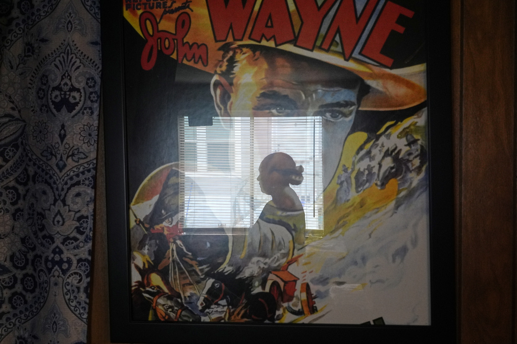 Paola Freites, who asked to be identified by her middle name and second last name to protect her family's safety, is reflected in a poster of John Wayne, who her teenaged son is a fan of, inside the two-bedroom mobile home where she lives with her husband and three children after fleeing persecution in their native Colombia, Thursday, Aug. 21, 2025, in Apopka, Fla. (AP Photo/Rebecca Blackwell)