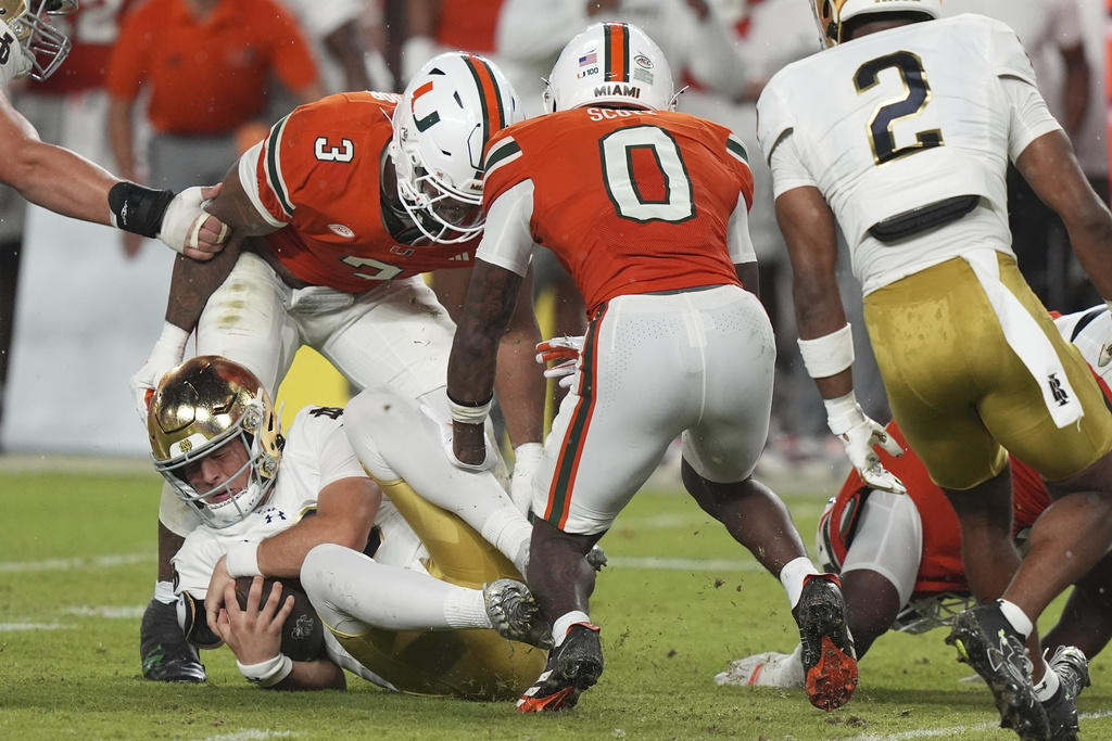 Notre Dame quarterback CJ Carr, left, falls to the field as Miami defensive lineman Akheem Mesidor (3) and defensive back Keionte Scott (0) defend during the first half of an NCAA college football game, Sunday, Aug. 31, 2025, in Miami Gardens, Fla. (AP Photo/Lynne Sladky)