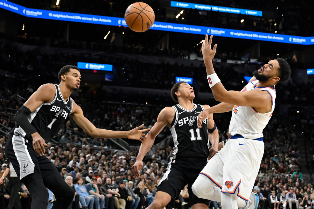 New York Knicks center Karl-Anthony Towns, right, is fouled as he drives against San Antonio Spurs forward Carter Bryant (11) and Spurs center Victor Wembanyama during the first half of an NBA basketball game, Wednesday, Dec. 31, 2025, in San Antonio. (AP Photo/Darren Abate)