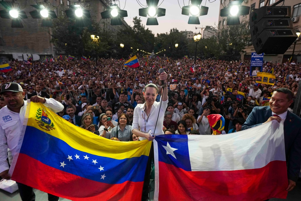 Venezuela's opposition leader Maria Corina Machado holds up a microphone next to Venezuelan and Chilean flags during a meeting with the Venezuelan community in Santiago, Chile, Thursday, March 12, 2026. (AP Photo/Esteban Felix)