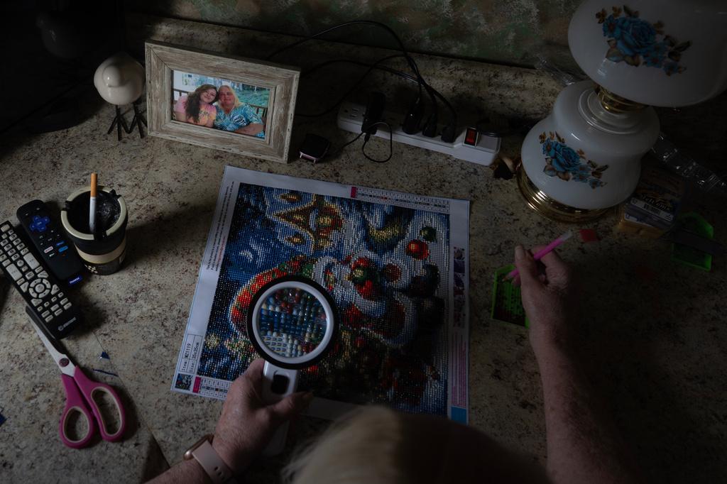 Rebecca Michalski works on her diamond painting kit by window light, in an effort to save money on her electricity bill, at her home in Rainelle, W.Va., Saturday, March 21, 2026, next to a photo of her daughter Sabrina Nicole Adkins, who passed away last year and whose funeral costs were difficult to afford. (AP Photo/Carolyn Kaster)