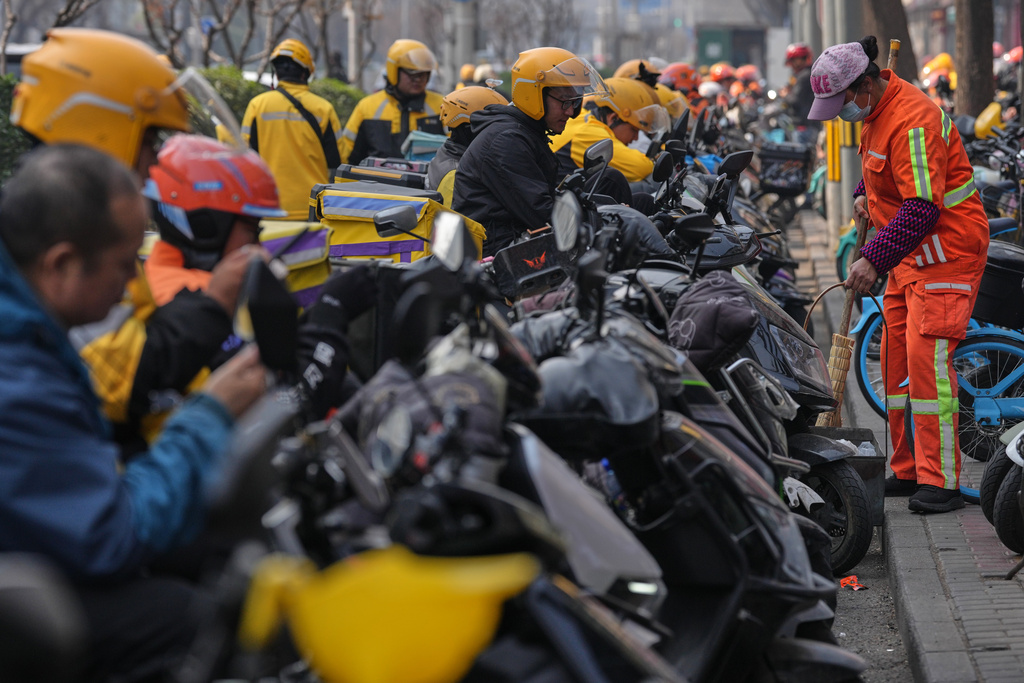 A cleaner sweeps near food delivery riders gather outside restaurants waiting for online orders, in Beijing, China, Wednesday, March 25, 2026. (AP Photo/Andy Wong)