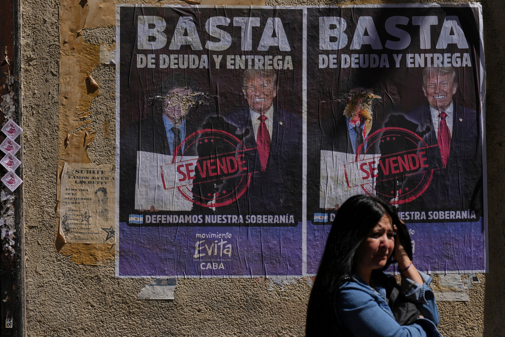 A woman waits for the bus in front of a poster with an image of President Javier Milei and President Donald Trump and a message that reads in Spanish; "Enough of debt and surrender'" in Buenos Aires, Argentina, Thursday, Oct. 2, 2025. (AP Photo/Natacha Pisarenko)