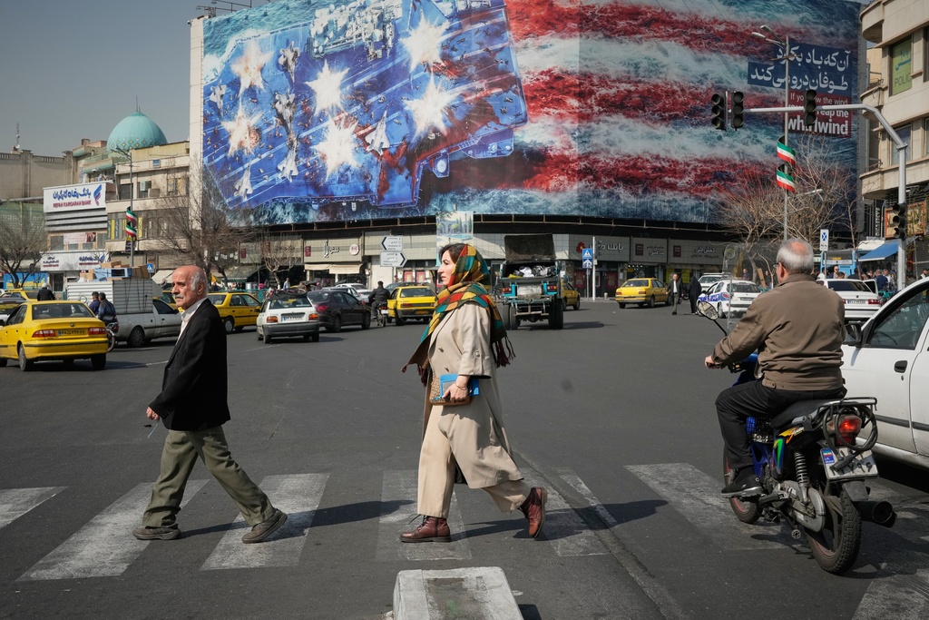 Pedestrians walk past a billboard depicting a U.S. aircraft carrier with damaged fighter jets on its deck and a sign in Farsi and English reading, "If you sow the wind, you'll reap the whirlwind," at Enqelab-e-Eslami (Islamic Revolution) Square in Tehran, Iran, Sunday, Feb. 22, 2026. (AP Photo/Vahid Salemi)