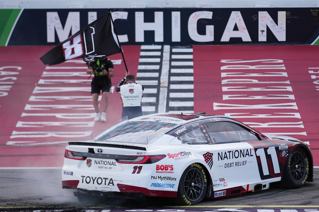 Denny Hamlin celebrates winning while honoring Kyle Busch with a flag after a NASCAR Cup Series auto race at Michigan International Speedway in Brooklyn, Mich., Sunday, June 7, 2026. (AP Photo/Paul Sancya)