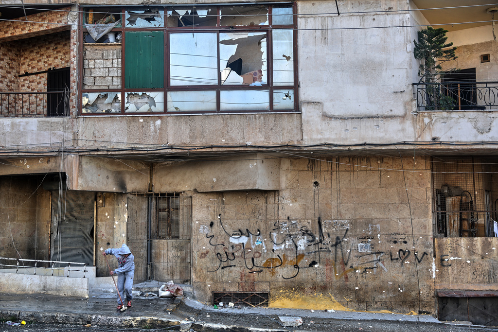 A Syrian girl sweeps debris along a street lined with damaged buildings in the Achrafieh neighborhood after a ceasefire was announced following three days of clashes between government forces and Kurdish fighters in a contested area of the northern city of Aleppo, Syria, Friday, Jan. 9, 2026. (AP Photo/Ghaith Alsayed)