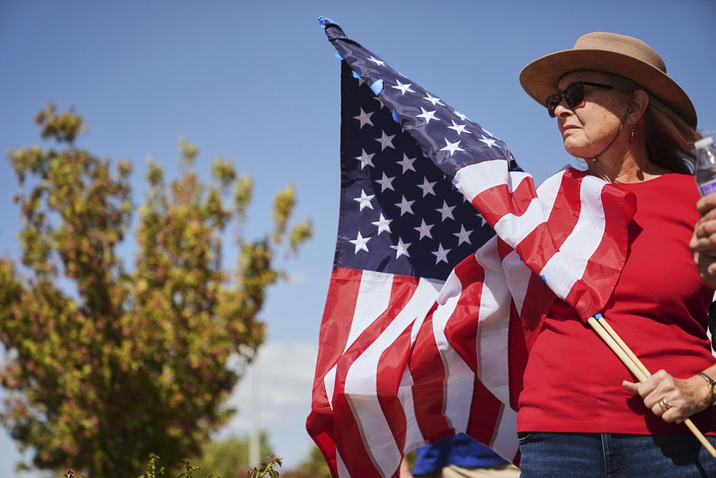A woman holding an American flag stands outside the entrance of the Roland R. Wright Air National Guard Base in Salt Lake City, Thursday, Sept. 11, 2025, where a hearse transporting the remains of Charlie Kirk arrived to be flown in Air Force Two to his home in Arizona. (AP Photo/Lindsey Wasson)