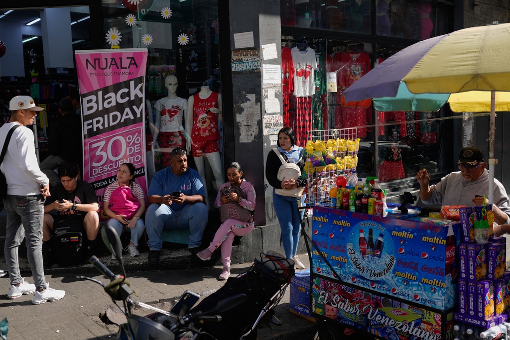 People sit outside a storefront promoting Black Friday discounts in Caracas, Venezuela, Friday, Nov. 28, 2025. (AP Photo/Ariana Cubillos)