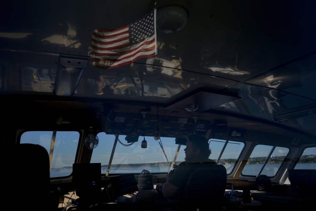 An American flag is reflected in the windows of the bridge as mate Peter Emery pilots the Gaspee, a crew transport vessel, along the Sakonnet River near Portsmouth, R.I., during the eight-hour trip to the Liftboat Robert platform and Expedition 501 in the North Atlantic, Saturday, July 19, 2025. (AP Photo/Carolyn Kaster)