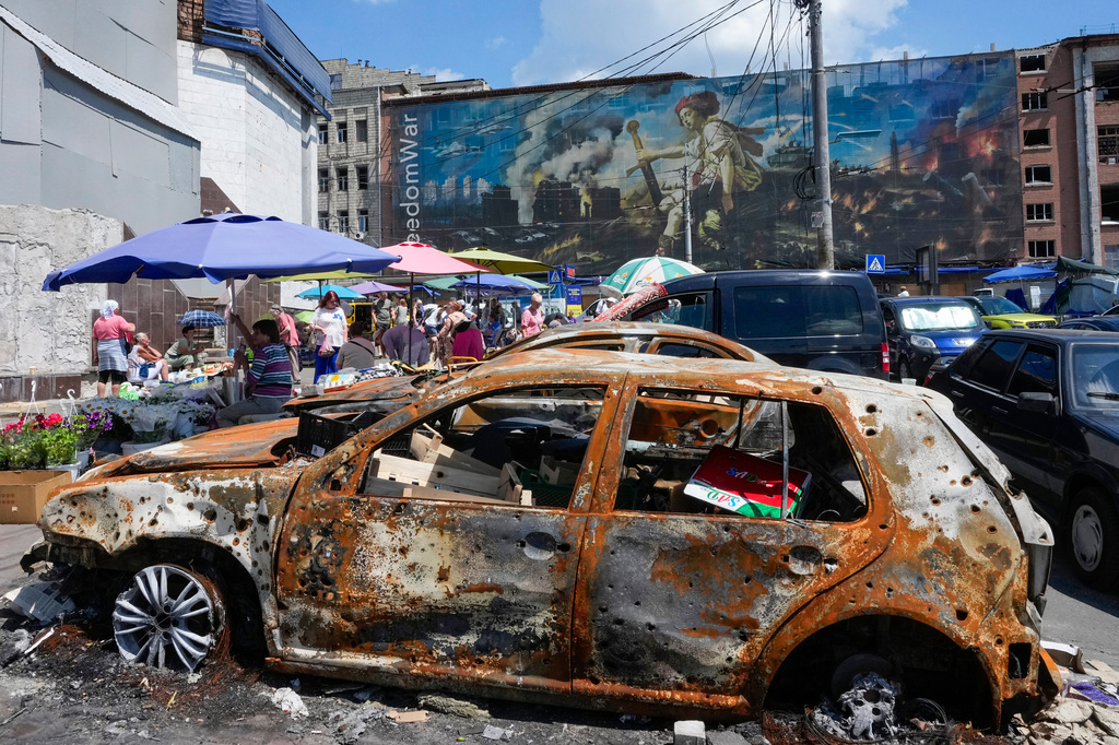 People buy food at an improvised outdoor market, burnt cars in the foreground, surrounded by damaged buildings covered with street artists paintings close to a big city marketplace that was ruined recently by Russian missiles in Kyiv, Ukraine, Monday, June 22, 2026. (AP Photo/Efrem Lukatsky)