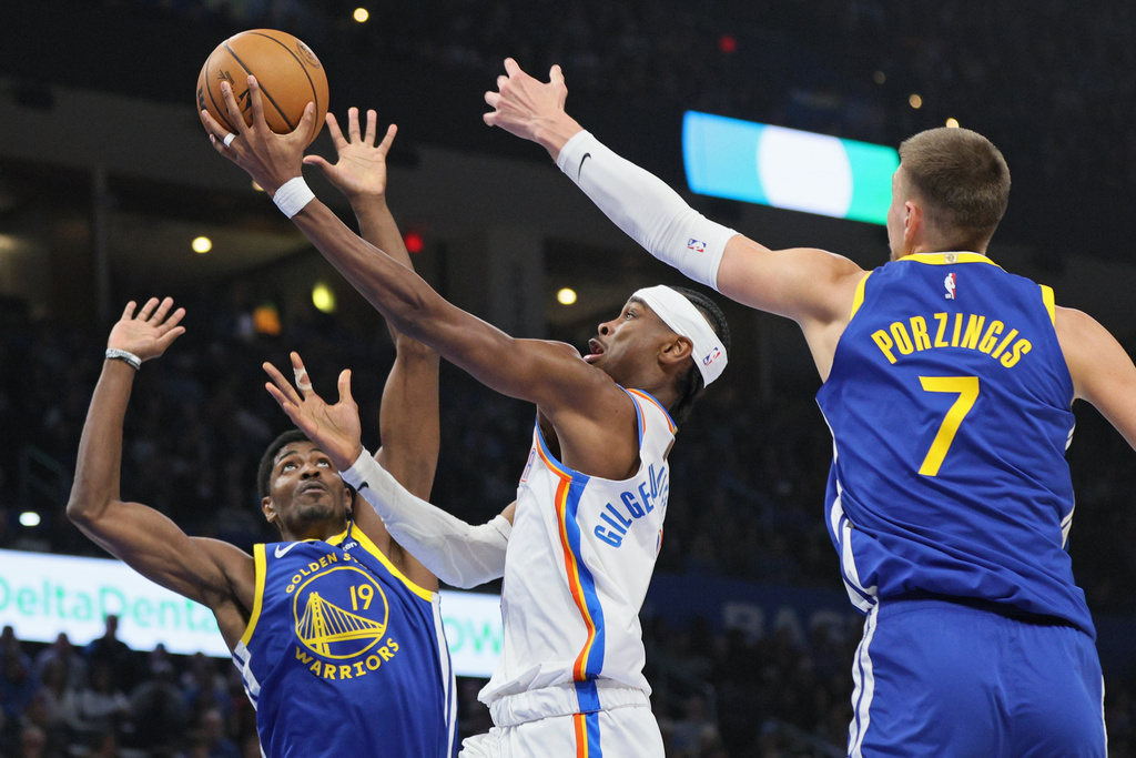 Oklahoma City Thunder guard Shai Gilgeous-Alexander, center, takes the ball to the basket between Golden State Warriors guard Nate Williams (19) and center Kristaps Porzingis (7) during the first half of an NBA basketball game Saturday, March 7, 2026, in Oklahoma City. (AP Photo/Nate Billings)