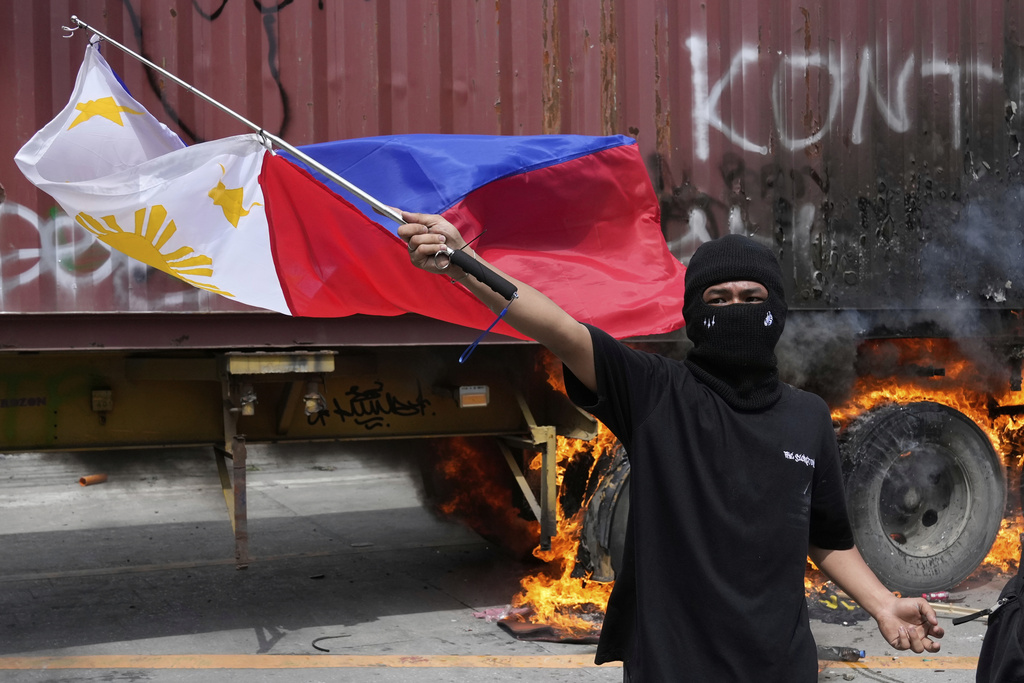 A protester waves a Philippine flag beside a burning container truck as they clash with police while trying to enter the Malacanang presidential palace compound in Manila, Philippines on Sunday, Sept. 21, 2025. (AP Photo/Aaron Favila)