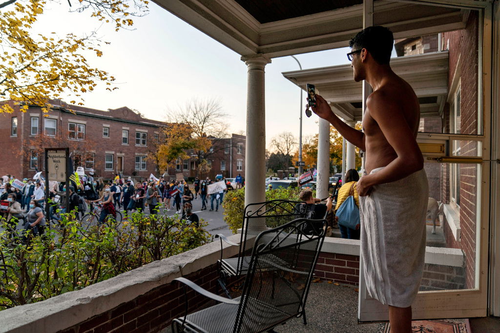 FILE - Karthik Mantha stands on his front porch wearing a towel, just as he was about to step into a shower, to watch a rally pass by celebrating the presidential election results in Detroit, Nov. 7, 2020. (AP Photo/David Goldman, File)