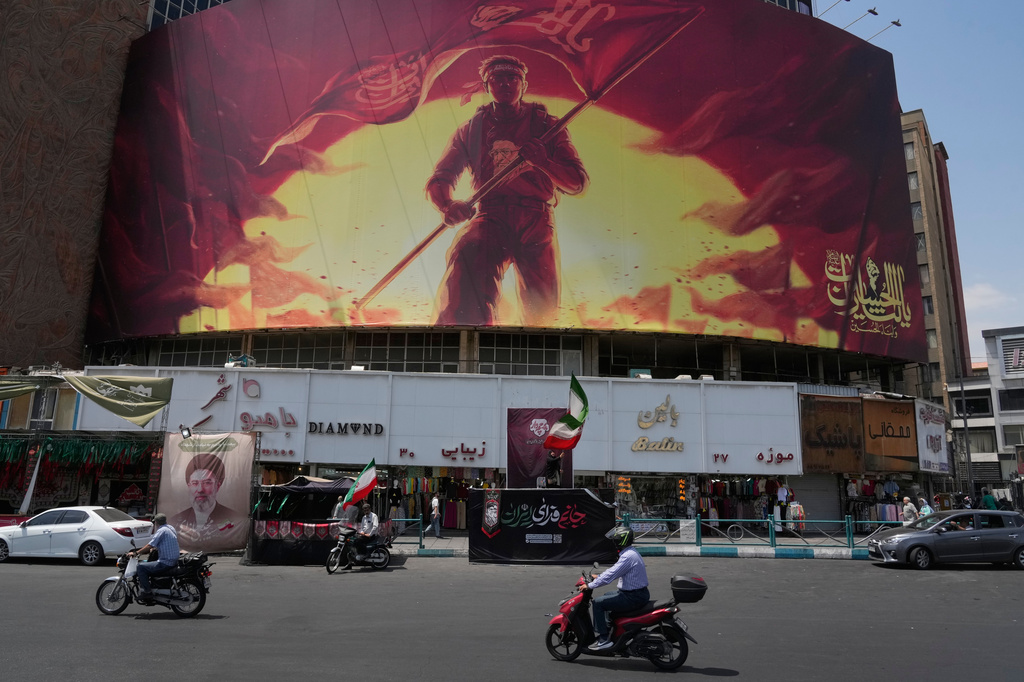 Motorbikes drive past a billboard showing a graphic calling for revenge on Imam Hussein, one of Shiite Islam's most beloved saints, who was killed in a 7th century battle, and the late Iranian Supreme Leader Ayatollah Ali Khamenei, who was killed during the U.S. and Israel strike on Feb. 28, in a square in downtown Tehran, Iran, Saturday, Aug. 1, 2026. (AP Photo/Vahid Salemi)