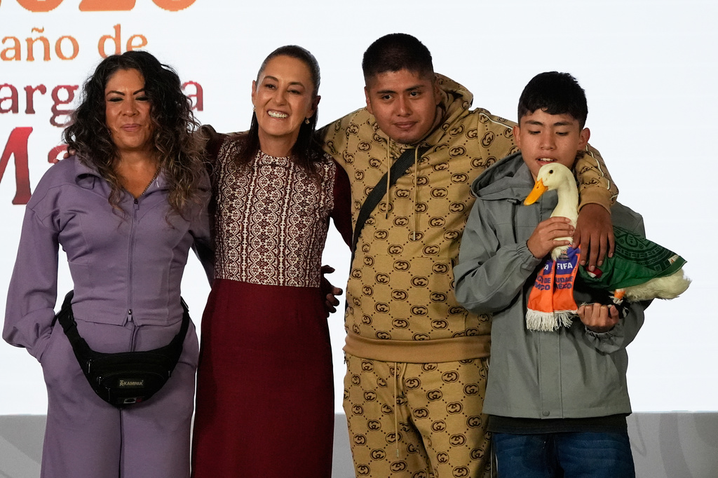 Merlin the duck, dressed in a Mexican national soccer team jersey, attends the daily press conference by Mexican President Claudia Sheinbaum, second from left, along with his caretakers Carla Gomez and her sons Carlos, and Christian who holds Merlin, at the National Palace in Mexico City, Monday, June 22, 2026. (AP Photo/Marco Ugarte)