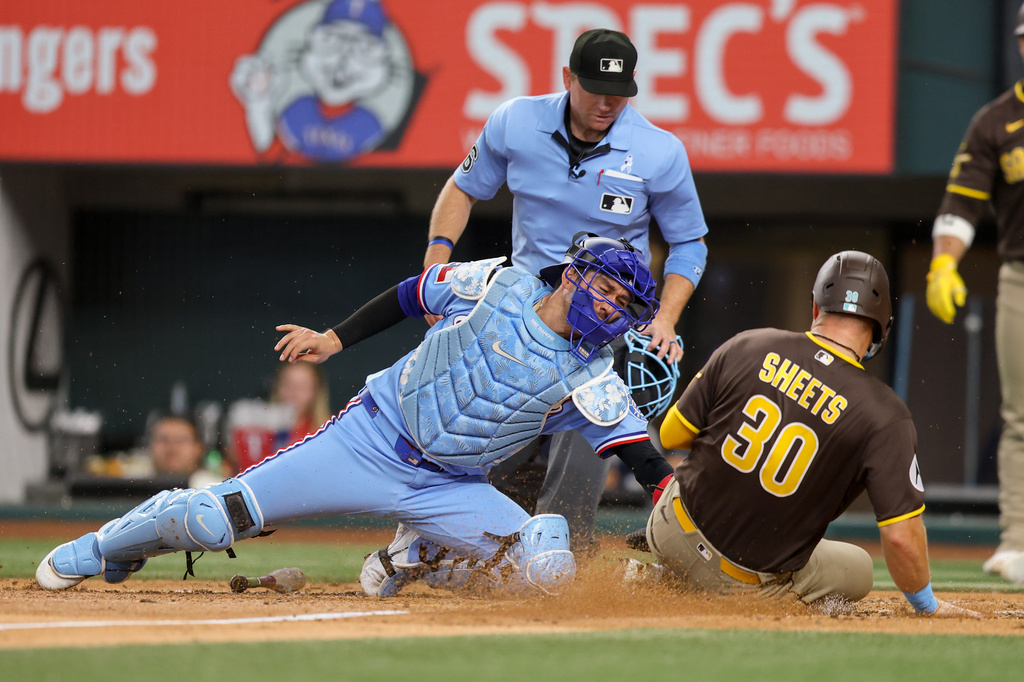 Texas Rangers catcher Kyle Higashioka, left, tags out San Diego Padres' Gavin Sheets (30) at the plate during the fourth inning of a baseball game, Sunday, June 21, 2026, in Arlington, Texas. (AP Photo/Gareth Patterson)