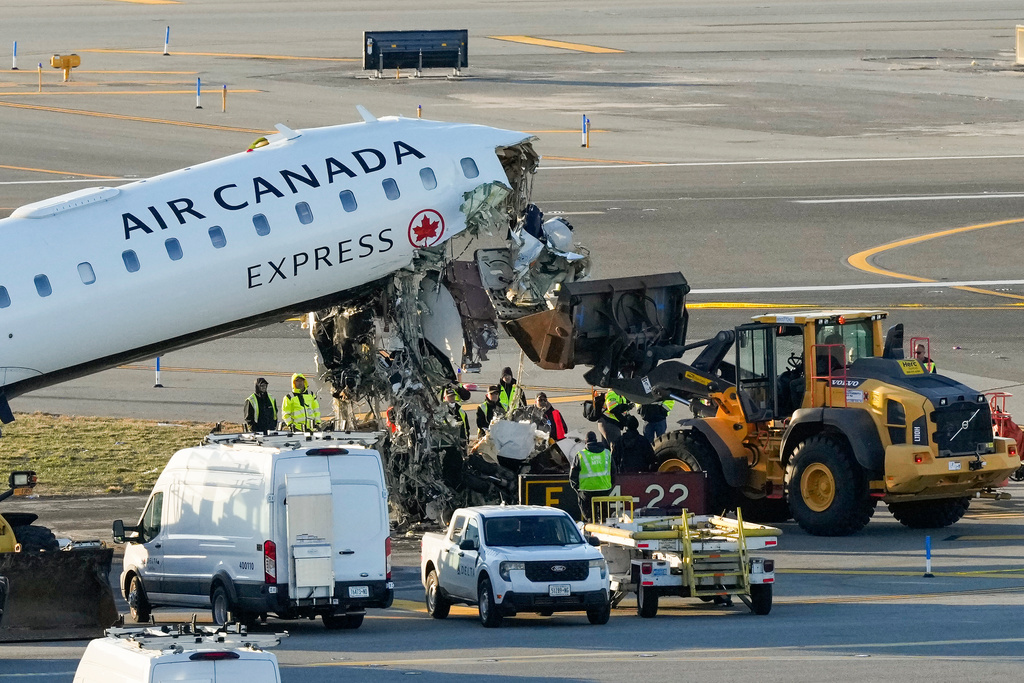 Aircraft maintenance workers position a front loader under debris hanging from the wreckage of an Air Canada Express jet, Tuesday, March 24, 2026, just off the runway where it had collided with a Port Authority fire truck Sunday night at LaGuardia Airport in New York. (AP Photo/Yuki Iwamura)