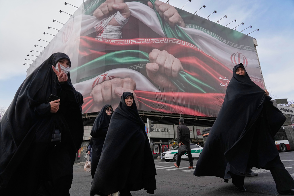FILE - Women, one flashing a victory hand gesture, cross a street under a huge banner showing hands firmly holding Iranian national flags as a sign of patriotism, in Tehran, Iran, Jan. 14, 2026. (AP Photo/Vahid Salemi, File)