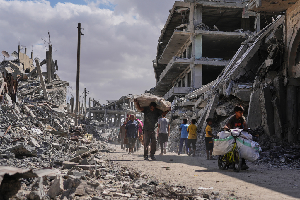 Displaced Palestinians walk with their belongings past destroyed buildings as they return to their homes in Khan Younis, southern Gaza Strip, Friday, Oct. 10, 2025, after Israel and Hamas agreed to a pause in their war and the release of the remaining hostages. (AP Photo/Jehad Alshrafi)