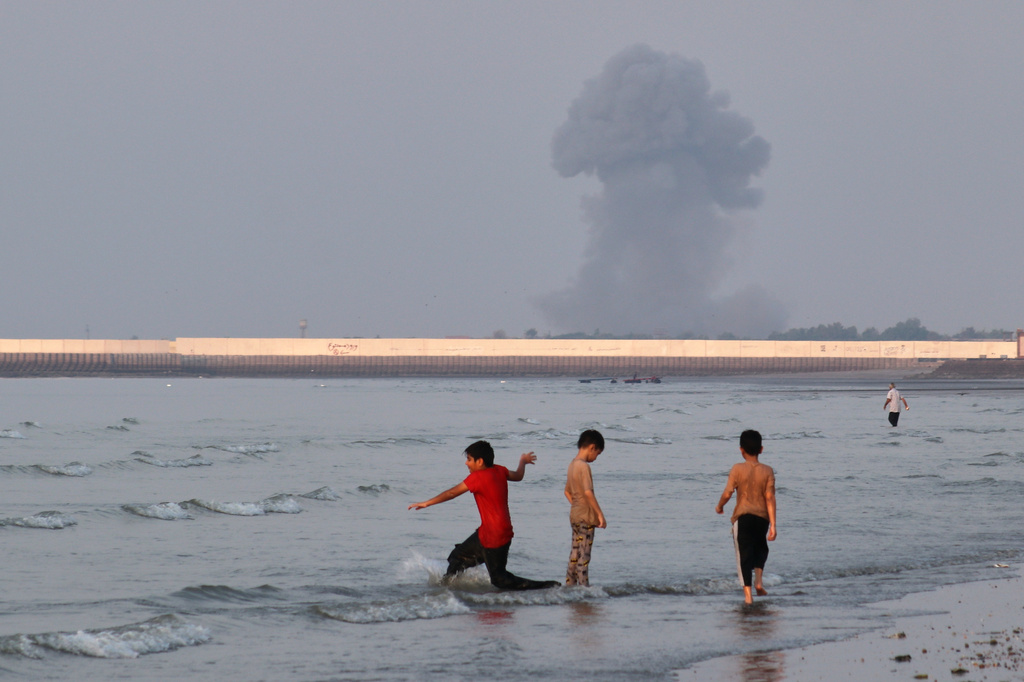 Three boys play in the shallow waters of the Strait of Hormuz, as a plume of smoke rises from an explosion in the background, off Bandar Abbas, Iran, Monday, July 13, 2026. (Razieh Poudat/ISNA via AP)