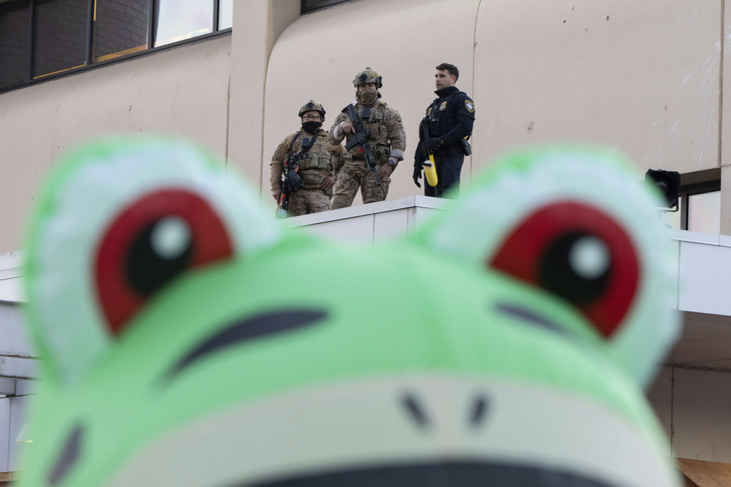 Law enforcement officers watch from a ledge of a United States Immigration and Customs Enforcement (ICE) facility as a protester stands outside in an inflatable frog costume on Tuesday, Oct. 21, 2025, in Portland, Ore. (AP Photo/Jenny Kane)