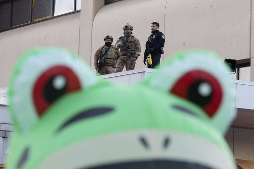 Law enforcement officers watch from a ledge of a United States Immigration and Customs Enforcement (ICE) facility as a protester stands outside in an inflatable frog costume on Tuesday, Oct. 21, 2025, in Portland, Ore. (AP Photo/Jenny Kane) Law enforcement officers watch from a ledge of a United States Immigration and Customs Enforcement (ICE) facility as a protester stands outside in an inflatable frog costume on Tuesday, Oct. 21, 2025, in Portland, Ore. (AP Photo/Jenny Kane)