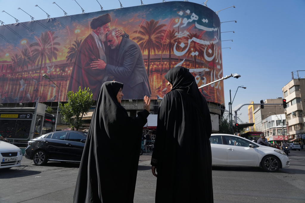 Women talk in front of a banner with graphic depicting the slain Iranian Supreme Leader Ayatollah Ali Khamenei kissing head of the late commander of Iran's Revolutionary Guard expeditionary Quds Force, Gen. Qassem Soleimani, who was killed in a U.S. drone attack in 2020 in Iraq, as they wait for the green light to cross an intersection in downtown Tehran, Iran, Monday, June 29, 2026. (AP Photo/Vahid Salemi)