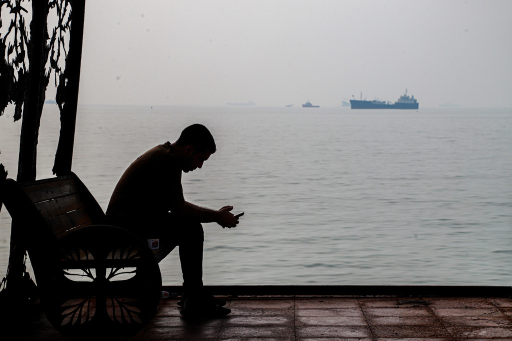A man checks his cellphone at a waterfront shelter as commercial vessels appear anchored in the Strait of Hormuz off Bandar Abbas, Iran, Monday, Tuesday, Oct, 6, 2026. (Amirhosein Khorgooi/ISNA via AP)