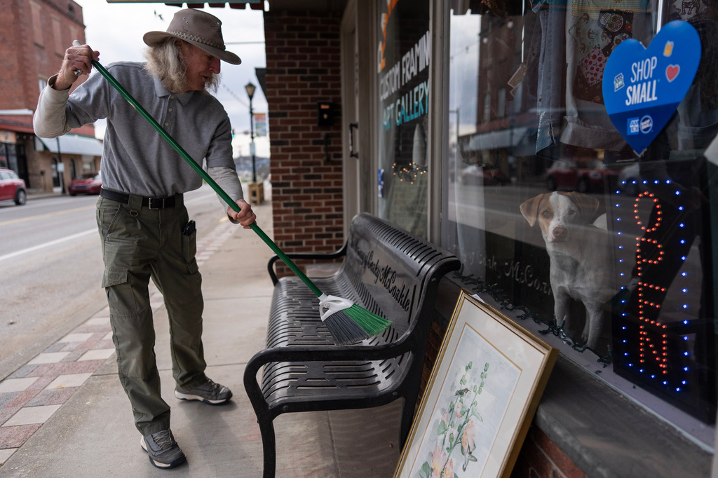 Philip Moulton sweeps the bench outside his Ravenswood Arts gallery where his dog JoJo peers through window, Friday, March 13, 2026, in Ravenswood W.Va., where surging electricity costs have forced some stores out of business. (AP Photo/Carolyn Kaster)