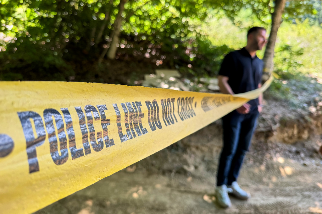 Erblin Ademi, right, whose father, Adem Ademi, a prominent surgeon, disappeared along with 22 other intellectuals during the Kosovo war in April 1999, stands behind yellow police tape near the excavation site of a mass grave in Small Kalluder, Kosovo, Wednesday, Aug. 5, 2026. (AP Photo/Zana Cimili)