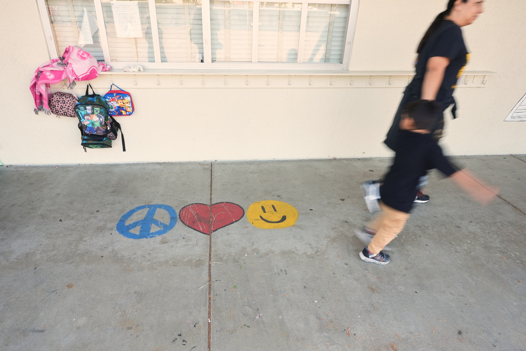 A teacher leads a child past a row of nearly empty hooks for backpacks outside a classroom at Perkins K-8 School Thursday, Nov. 13, 2025, in San Diego. (AP Photo/Gregory Bull)