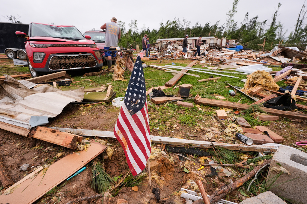 An American flag is seen as people walk among debris at Gene's Mobile Home Supply, a trailer park in Bogue Chitto, Miss., Thursday, May, 7, 2026, after a tornado cut across the state. (AP Photo/Gerald Herbert)
