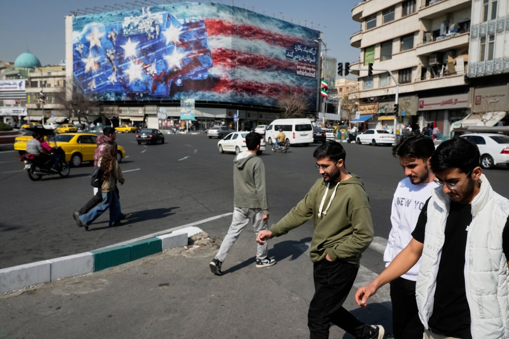 Pedestrians walk past a billboard depicting a U.S. aircraft carrier with damaged fighter jets on its deck and a sign in Farsi and English reading, "If you sow the wind, you'll reap the whirlwind," at Enqelab-e-Eslami (Islamic Revolution) Square in Tehran, Iran, Sunday, Feb. 22, 2026. (AP Photo/Vahid Salemi)
