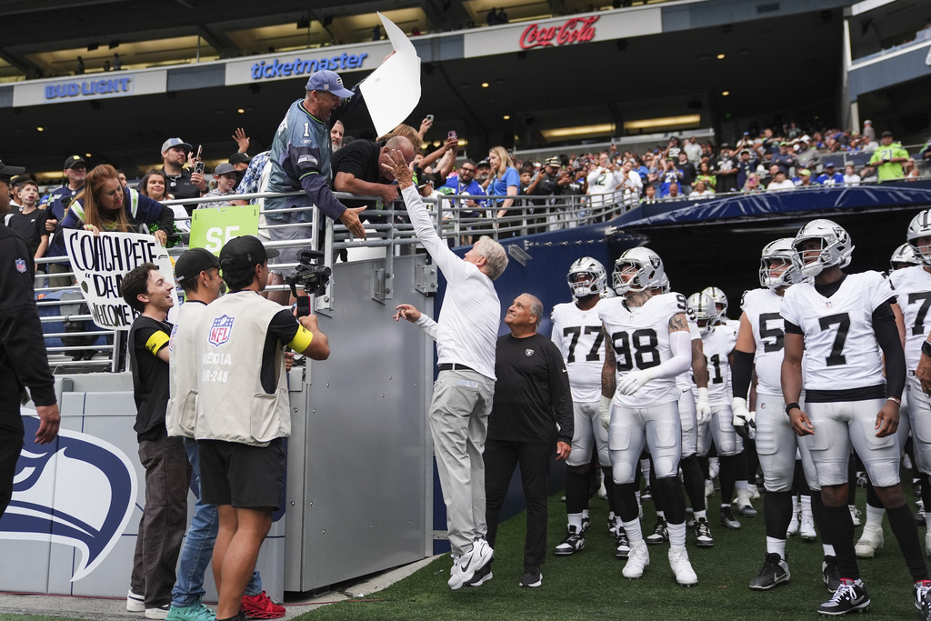 Las Vegas Raiders head coach Pete Carroll, center, tries to grab a sign that reads "Bigger Bust-Geno or JaMarcus Russell?" before an NFL preseason football game against the Seattle Seahawks, Thursday, Aug. 7, 2025, in Seattle. (AP Photo/Lindsey Wasson)