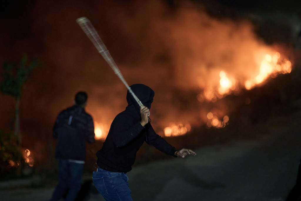 A Palestinian uses a slingshot during clashes with Israeli settlers as brush burns after it was set ablaze to prevent them from approaching the West Bank village of Sinjil, Friday, July 17, 2026. (AP Photo/Leo Correa)