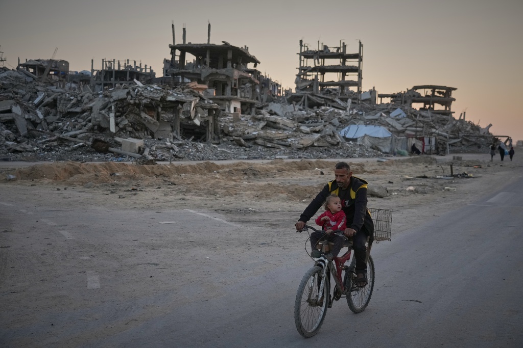 A man carries a child while riding his bicycle along a damaged street in the Al-Shati camp in Gaza City, Tuesday, Nov. 18, 2025. (AP Photo/Jehad Alshrafi)