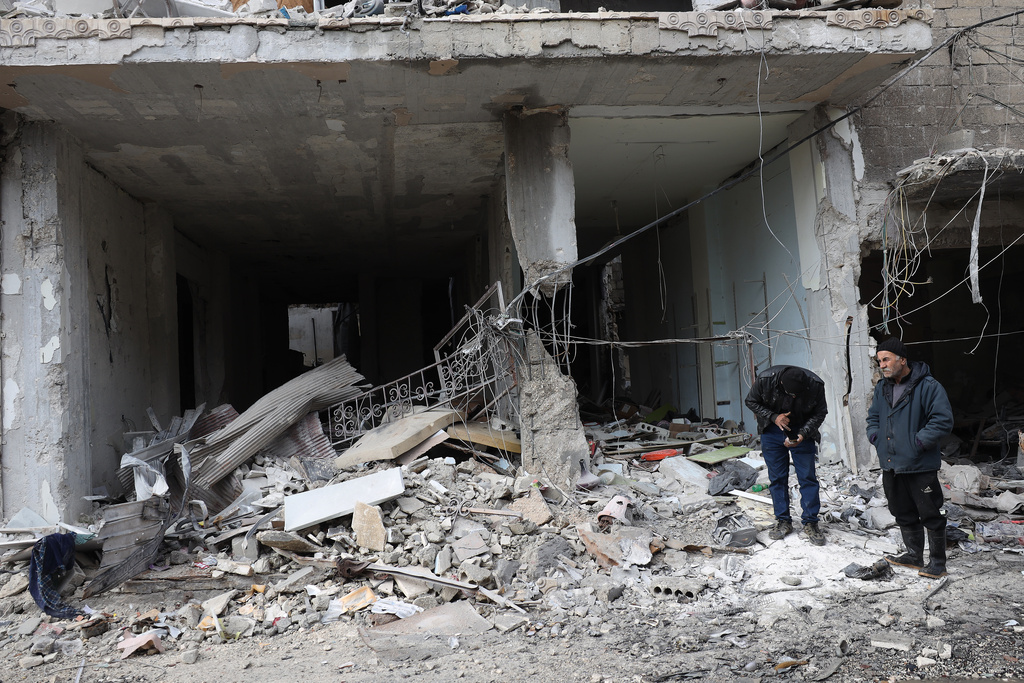 People stand in front of destroyed shops, in the Sheikh Maqsoud neighborhood after days of fighting between government forces and Kurdish fighters in the northern city of Aleppo, Syria, Tuesday, Jan. 13, 2026. (AP Photo/Omar Albam)