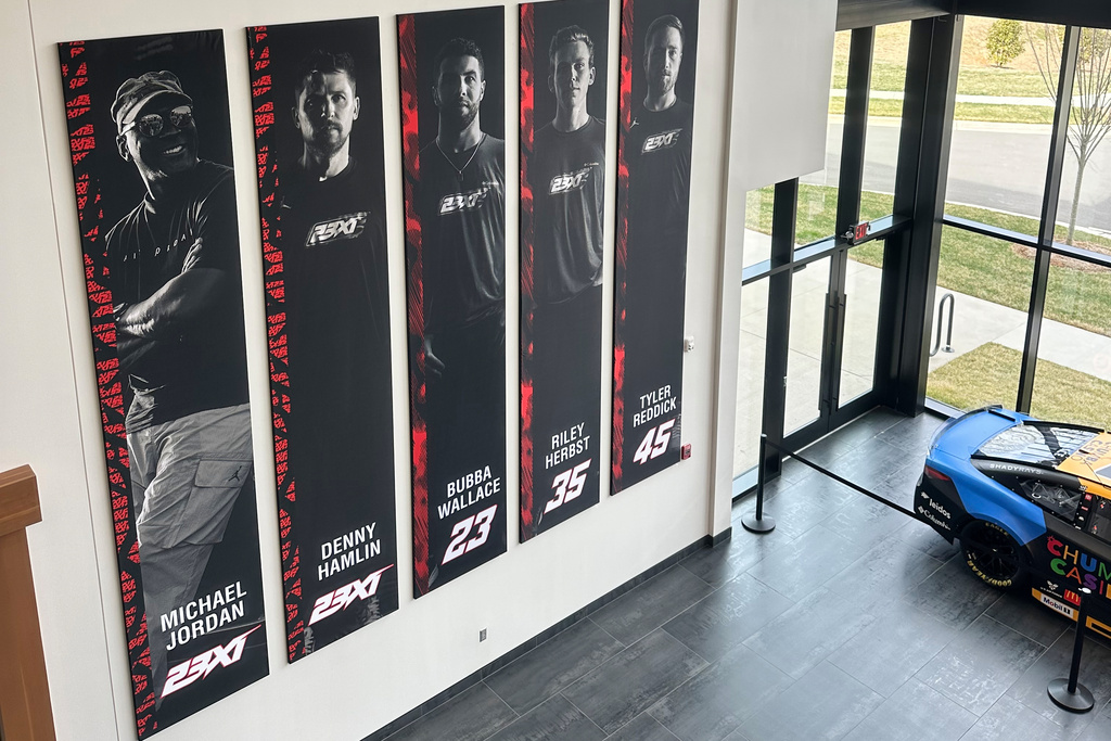A banner, showing co-owners Michael Jordan and Denny Hamlin, as well as drivers Bubba Wallace, Corey Heim and Tyler Reddick, hangs in the lobby at Airspeed, the headquarters of 23XI Racing, Tuesday, Feb. 24, 2026 in Huntersville, N.C. (AP Photo/Jenna Fryer)