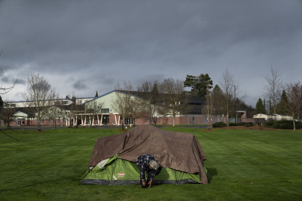 FILE - With Fruitdale Elementary School in the background, a homeless man adjusts his shoe at Fruitdale Park, March 23, 2024, in Grants Pass, Ore. (AP Photo/Jenny Kane, File)