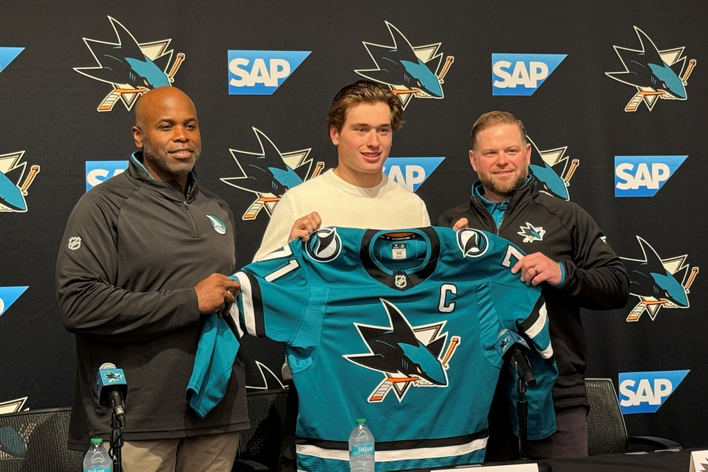 In this photo taken with a smartphone, San Jose Sharks forward Macklin Celebrini, center, is presented with the captain's jersey by general manager Mike Grier, left, and coach Ryan Warsofsky during an NHL hockey news conference in San Jose, Calif., Friday, Sept. 25, 2026. (AP Photo/Josh Dubow)