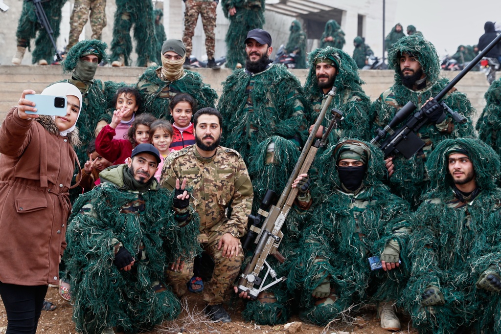 A child takes a selfie with soldiers of the new Syrian army dressed in camouflage as they gather before a parade, part of celebrations marking the first anniversary of the ousting of the Bashar Assad regime in Douma, on the outskirts of Damascus, Syria, Monday, Dec. 8, 2025. (AP Photo/Omar Sanadiki)