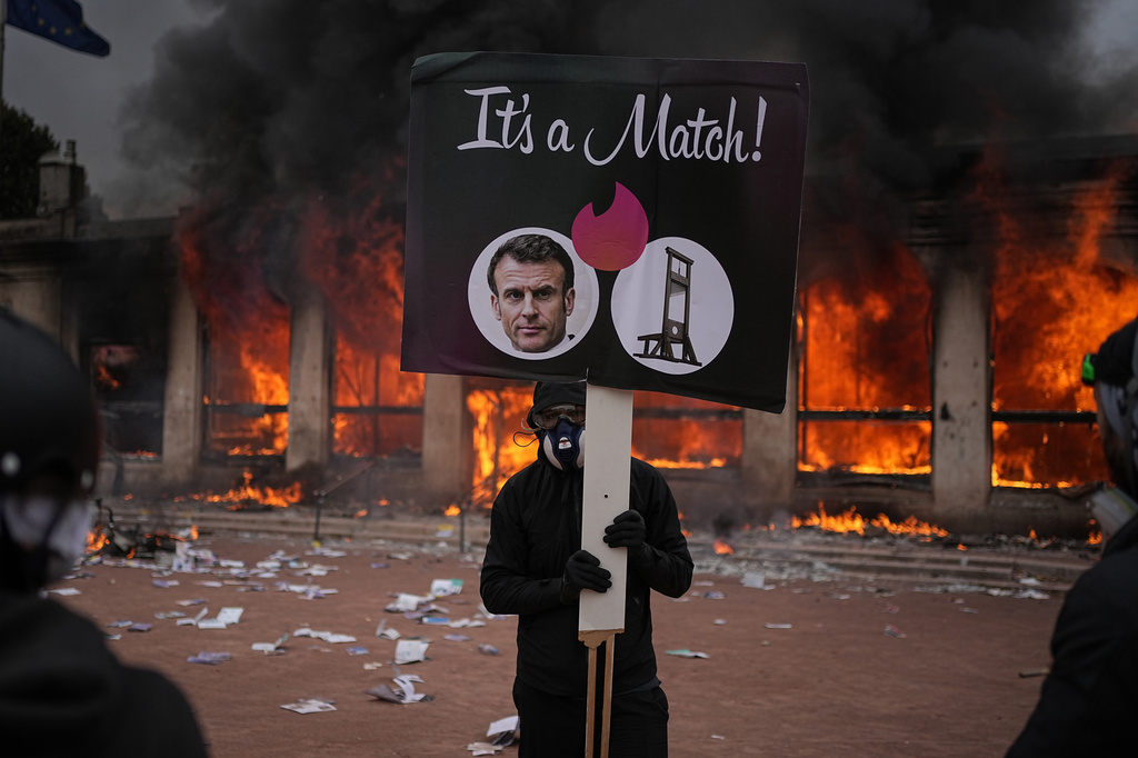 A protester holds a placard with a photo of French president Emmanuel Macron and a guillotine in front of a burning tourism office during a demonstration against rising living costs and in solidarity with students' grievances in Lyon, France, Saturday, Oct. 17, 2026. (AP Photo/Laurent Cipriani)